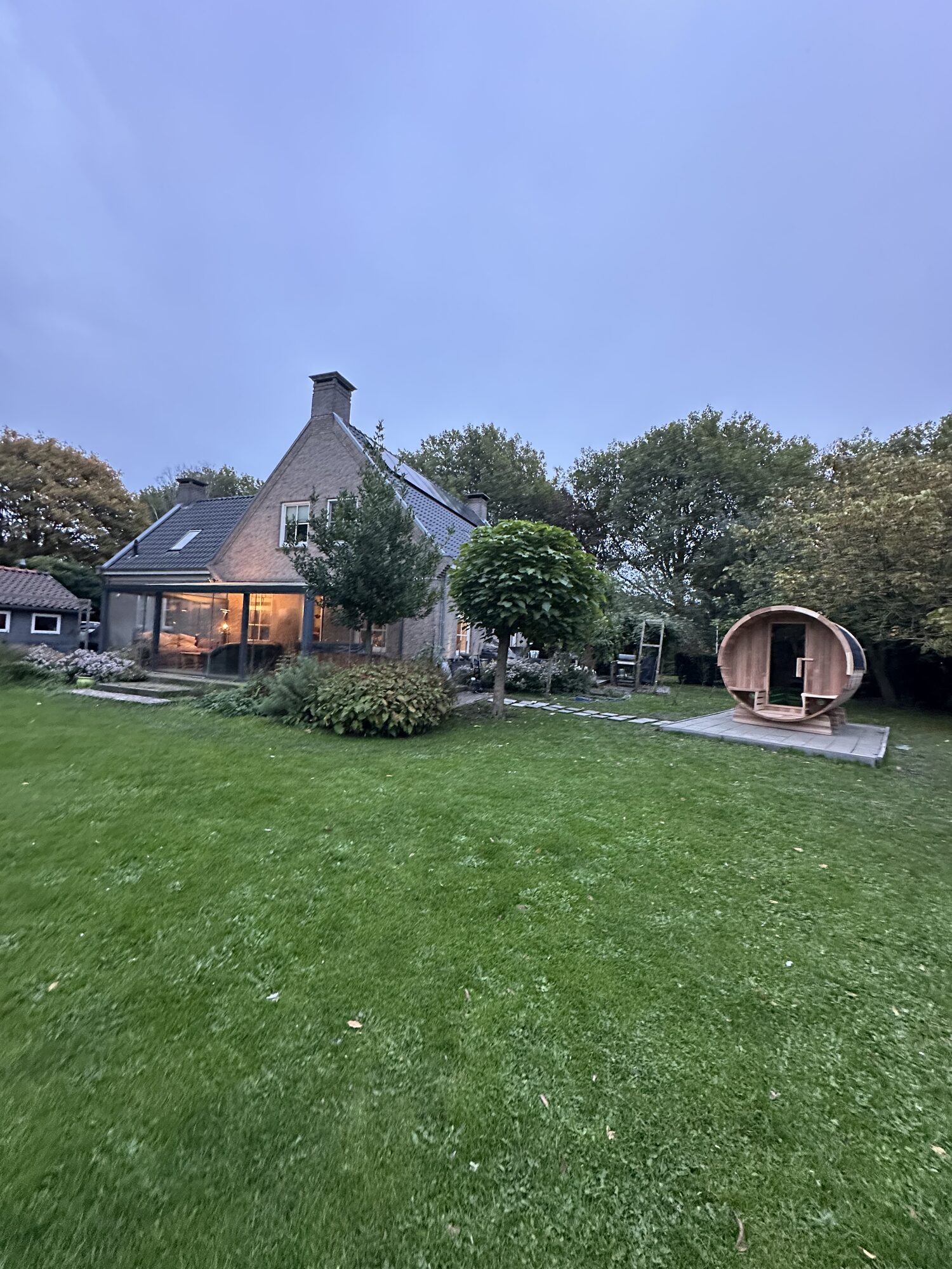 The villa in Aalsmeer at dusk with its lit glass veranda, large lawn and barrel sauna in the garden